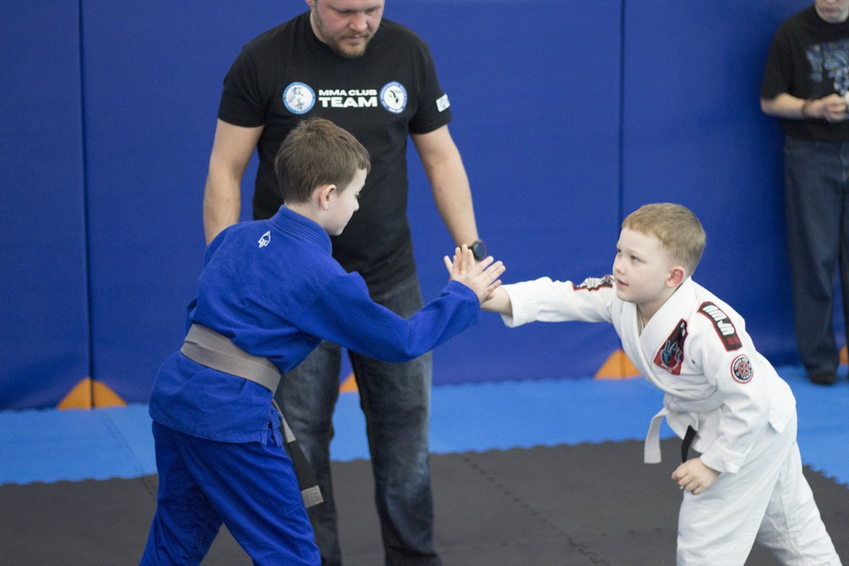 Dwaj chłopcy w kimonach witają się przed walką, między nimi sędzia w koszulce MMA Club Team