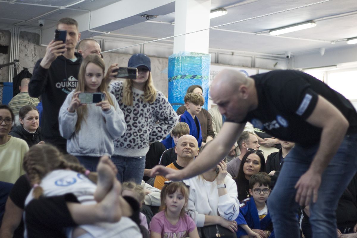 Sędzia pochyla się nad walczącymi dziećmi, w tle publiczność nagrywa walkę telefonami