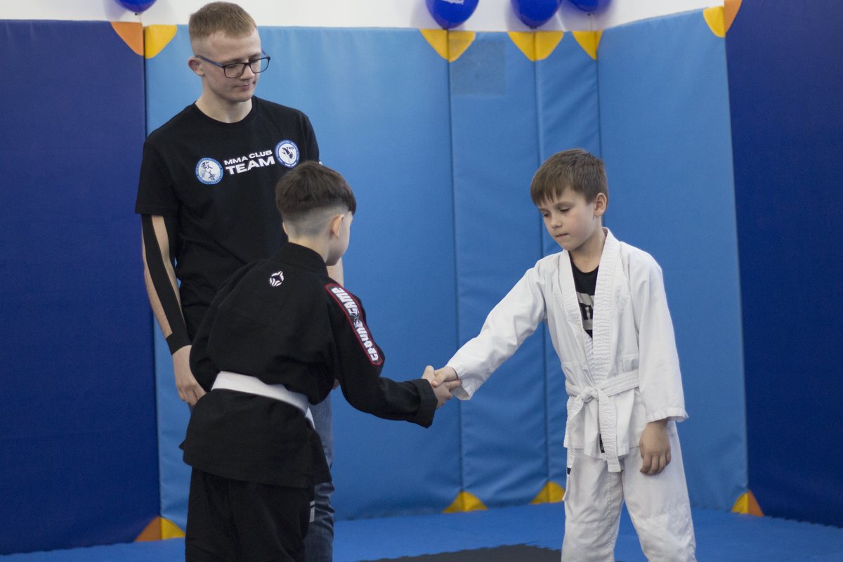 Dwaj chłopcy w kimonach podają sobie ręce na macie, obok prowadzący w koszulce MMA Club Team